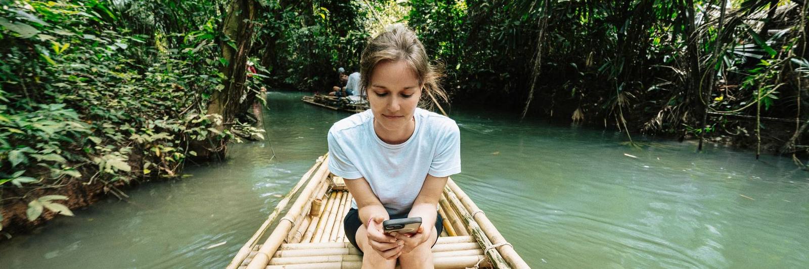 A traveller checking her phone on a bamboo raft on a jungle river in Thailand